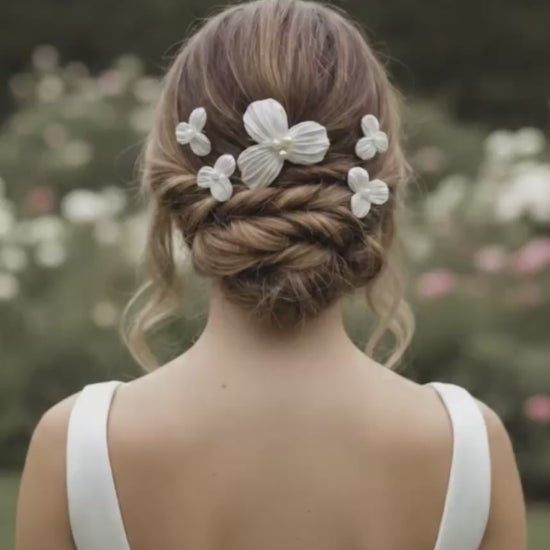 Video de novia de espaldas mostrando un peinado recogido trenzado decorado con horquillas de flores Dalia y sosteniendo un ramo de flores blancas.