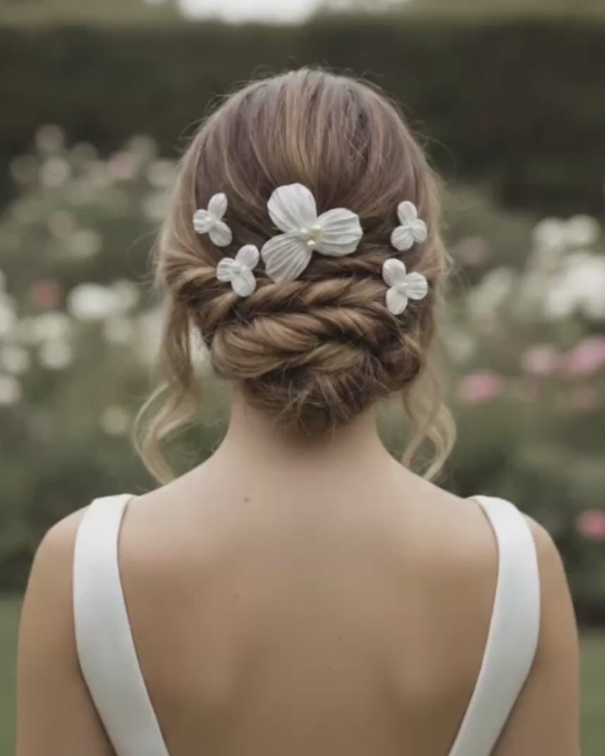 Video de novia de espaldas mostrando un peinado recogido trenzado decorado con horquillas de flores Dalia y sosteniendo un ramo de flores blancas.