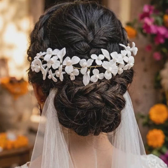 Video corto de 8 segundos que muestra un plano detalle trasero del peinado de una novia. Luce un recogido bajo trenzado texturizado adornado con una guía flexible de flores blancas de porcelana fría y perlas artesanales de Pedra México, de la cual cae un velo de novia de tul liso. El fondo es un jardín de bodas con flores borrosas
