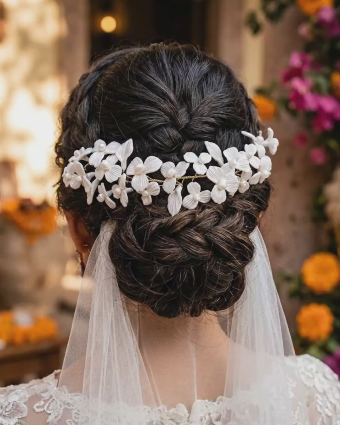 Video corto de 8 segundos que muestra un plano detalle trasero del peinado de una novia. Luce un recogido bajo trenzado texturizado adornado con una guía flexible de flores blancas de porcelana fría y perlas artesanales de Pedra México, de la cual cae un velo de novia de tul liso. El fondo es un jardín de bodas con flores borrosas