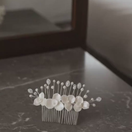 Video de producto de una peineta de novia Pedra en porcelana fría marfil. El accesorio reposa sobre una mesa de mármol, mostrando el detalle de las flores artesanales, perlas y la peineta plateada de sujeción