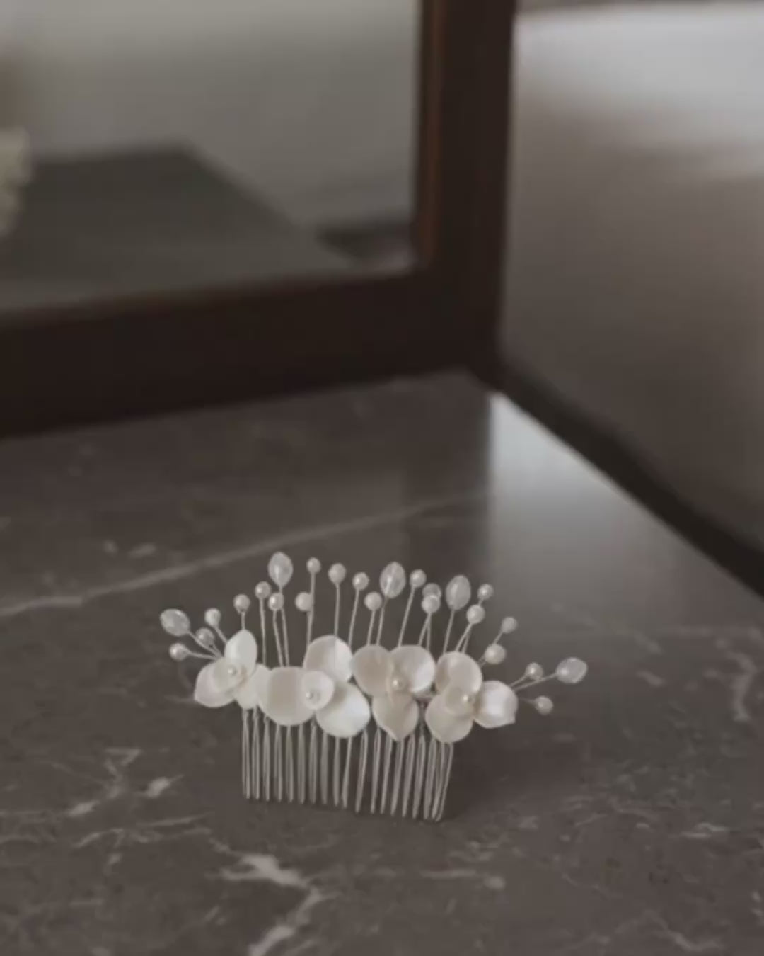 Video de producto de una peineta de novia Pedra en porcelana fría marfil. El accesorio reposa sobre una mesa de mármol, mostrando el detalle de las flores artesanales, perlas y la peineta plateada de sujeción