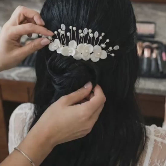 Primer plano de un tocado para novia Pedra hecho a mano con flores de porcelana marfil y detalles en perlas. Se muestra la textura y el acabado del accesorio siendo ajustado en un peinado de novia con cabello oscuro.